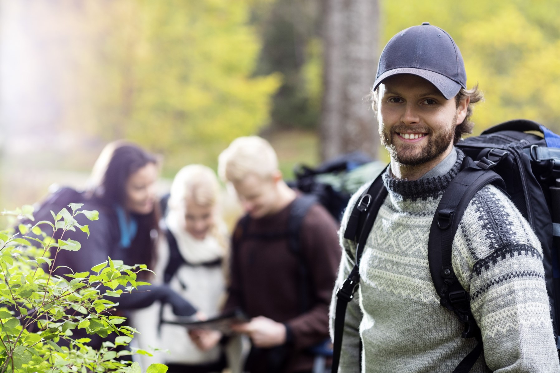 En yngre man i ylletröja och med ryggsäck på ryggen står ute i skogen, i bakgrunden en grupp som studerar en karta